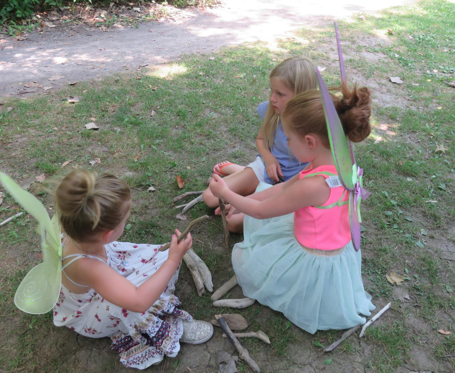 three young girls sitting on the ground in their fairy costumes working with sticks 